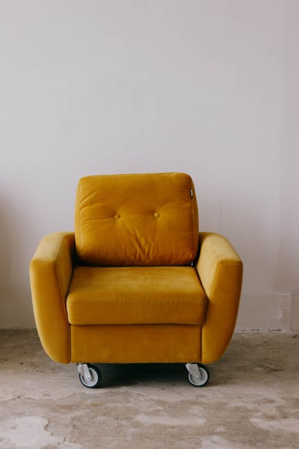 A mustard yellow armchair with a plush, tufted backrest and armrests, positioned against a plain white wall in a minimalist room. The chair is on small grey caster wheels, making it easily mobile. The