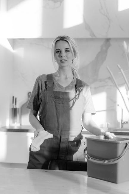 An interior scene of a modern, minimalist kitchen with white walls, light grey tiled flooring, and a large glass sliding door allowing natural light to fill the space. Three cleaning professionals, dr