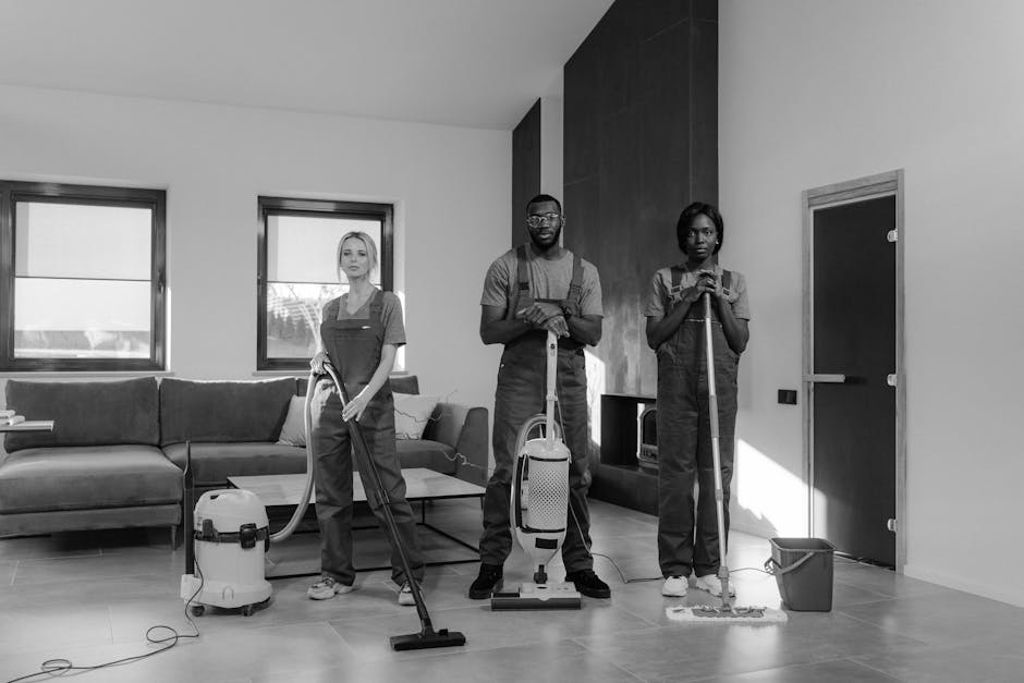 A team of three professional cleaners from Arnos Grove Cleaners is performing surface cleaning and deep cleaning in a modern living room with a minimalist design. The room features a light-colored woo
